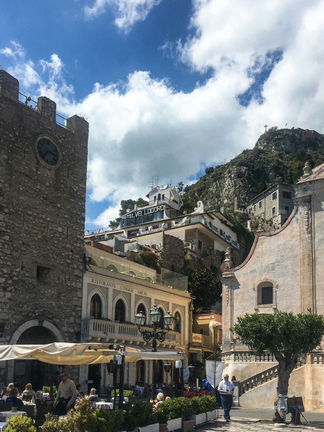 Una vista di una piazza europea con una prominente torre dell'orologio in pietra e edifici che costeggiano un ripido pendio verde sotto un cielo nuvoloso.