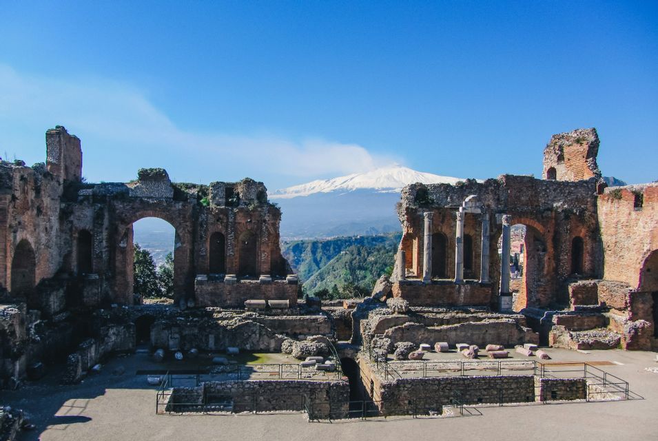 Le rovine in pietra di un antico anfiteatro con un vulcano innevato e fumante visibile sullo sfondo.