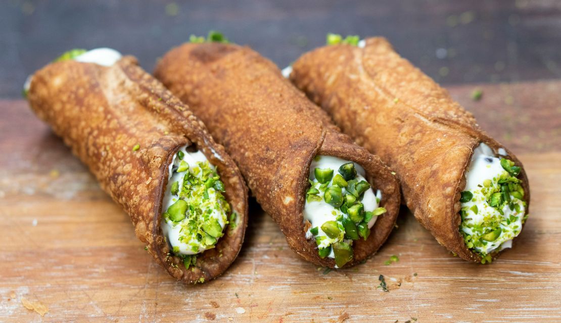 Primo piano di tre cannoli con ripieno di crema bianca, guarniti con pistacchi tritati su un tagliere di legno.