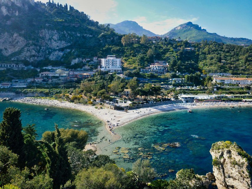 Una stretta striscia di sabbia si estende in una baia turchese, con una città arroccata su una lussureggiante collina verde sullo sfondo.