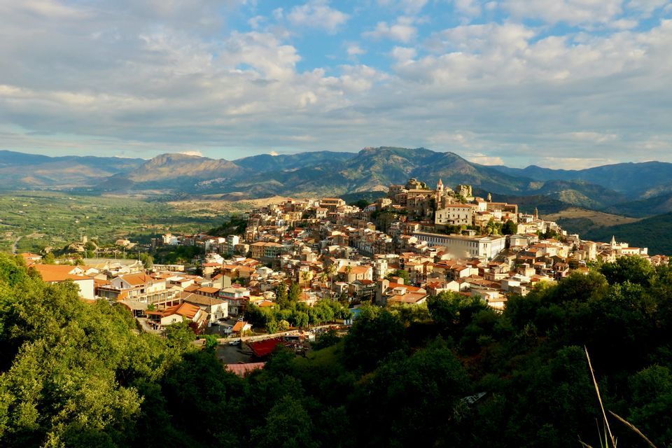 Una cittadina dai tetti in terracotta si adagia su un lussureggiante fianco verde, con una valle e una catena montuosa sullo sfondo sotto un cielo blu e nuvoloso.