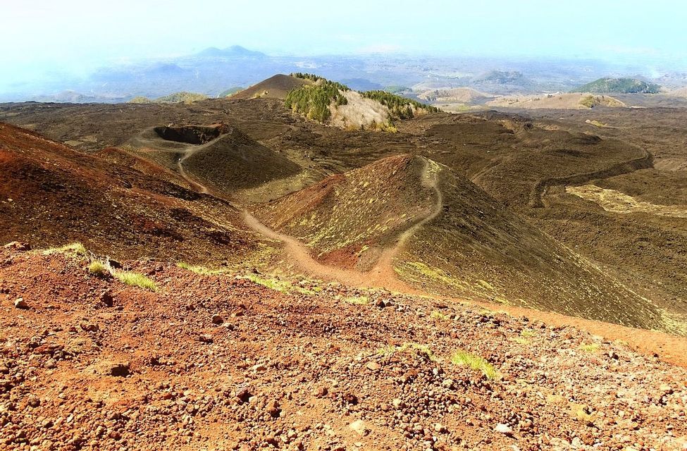 Un vasto paesaggio vulcanico con terra rossastra, piccoli crateri e un sentiero sterrato serpeggiante sotto un cielo azzurro chiaro