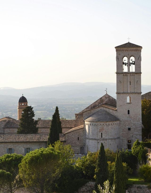 Un complesso di chiese in pietra, con un alto campanile e tetti in tegole, immerso tra alberi verdi e affacciato su una valle nebbiosa.