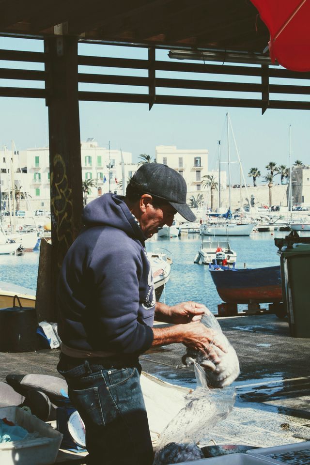 Un pescatore con felpa blu e cappello maneggia un grosso pesce su un molo, con un porto soleggiato pieno di barche sullo sfondo.