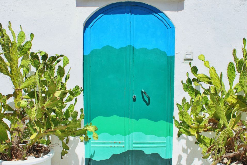 Una porta ad arco dipinta nelle tonalità del blu e del verde è incastonata in una parete bianca, affiancata da due vasi di fichi d'India.