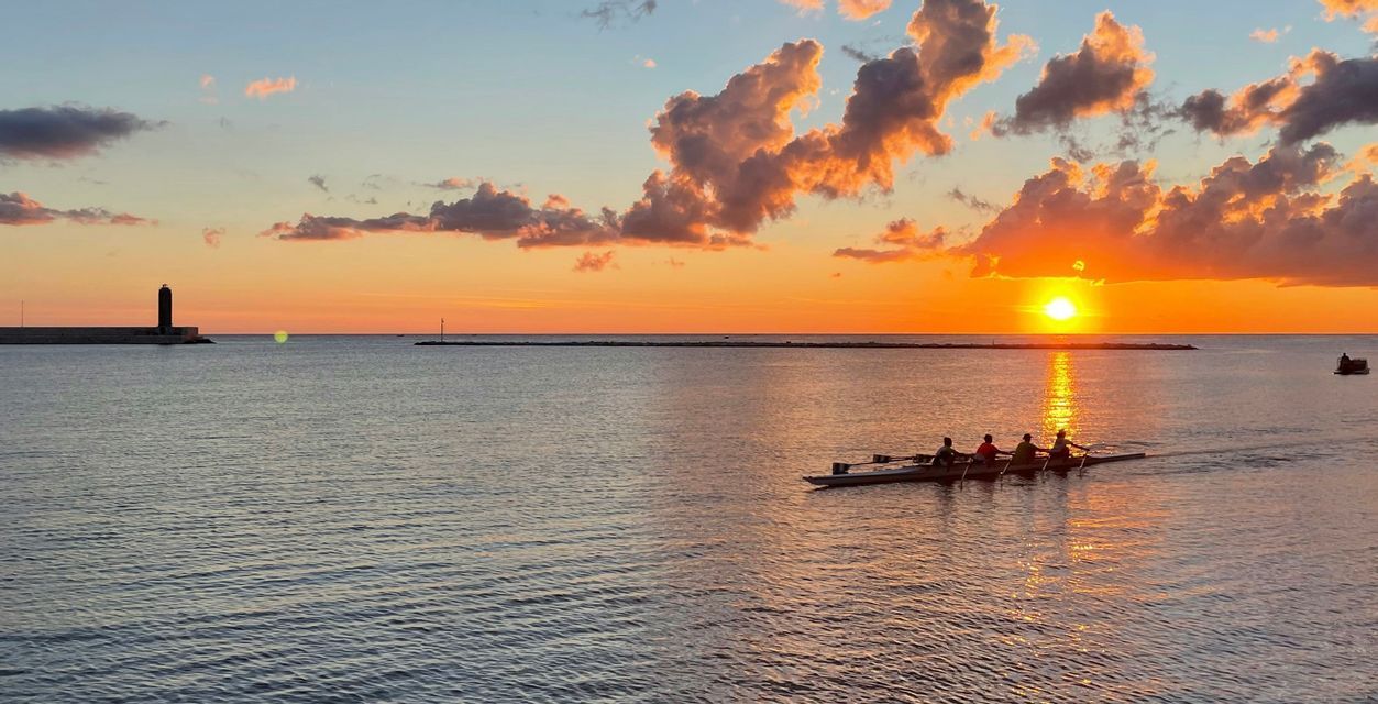 Quattro persone su una barca a remi su acqua calma al tramonto, con un faro su un molo sullo sfondo.