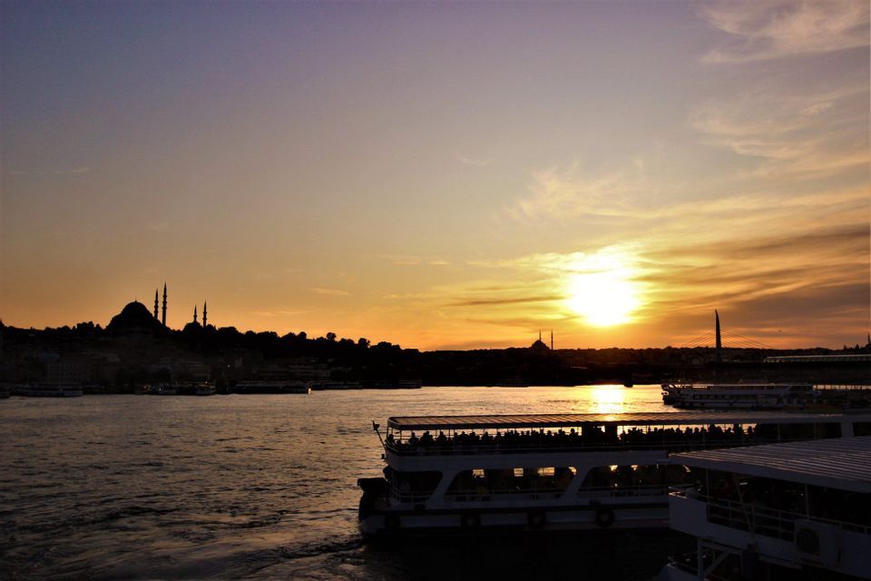 Ferries con pasajeros navegan sobre el agua al atardecer, con el perfil de la ciudad y mezquitas en silueta al fondo.