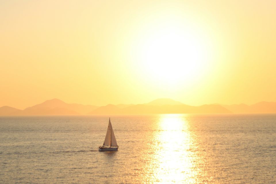 Una barca a vela solitaria sul mare durante un tramonto dorato, con montagne lontane all'orizzonte