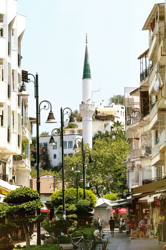 Un minareto bianco con una cima conica verde si erge sopra una stretta via cittadina fiancheggiata da alberi, edifici e lampioni ornati.
