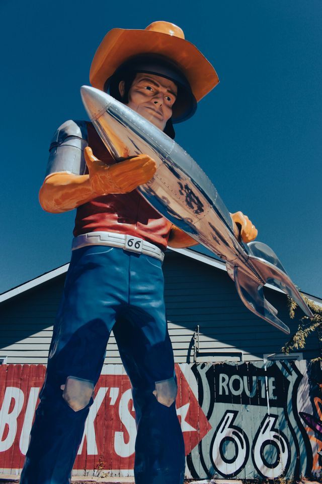 Una gran estatua de carretera de un hombre con sombrero de vaquero sosteniendo un cohete plateado frente a un letrero de la Ruta 66.