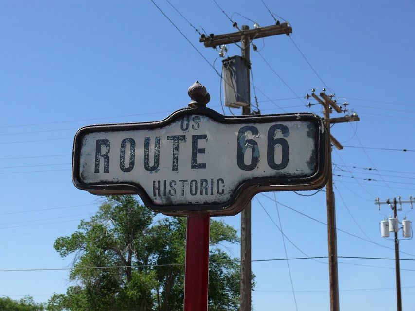 Primer plano de un cartel desgastado de la Ruta Histórica 66 de EE. UU. en un poste rojo, con un cielo azul y postes de servicios de fondo.