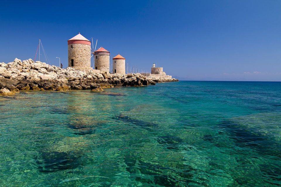 Tre mulini a vento in pietra con tetti conici rossi si ergono su una costa rocciosa accanto a un mare cristallino turchese sotto un cielo blu profondo.