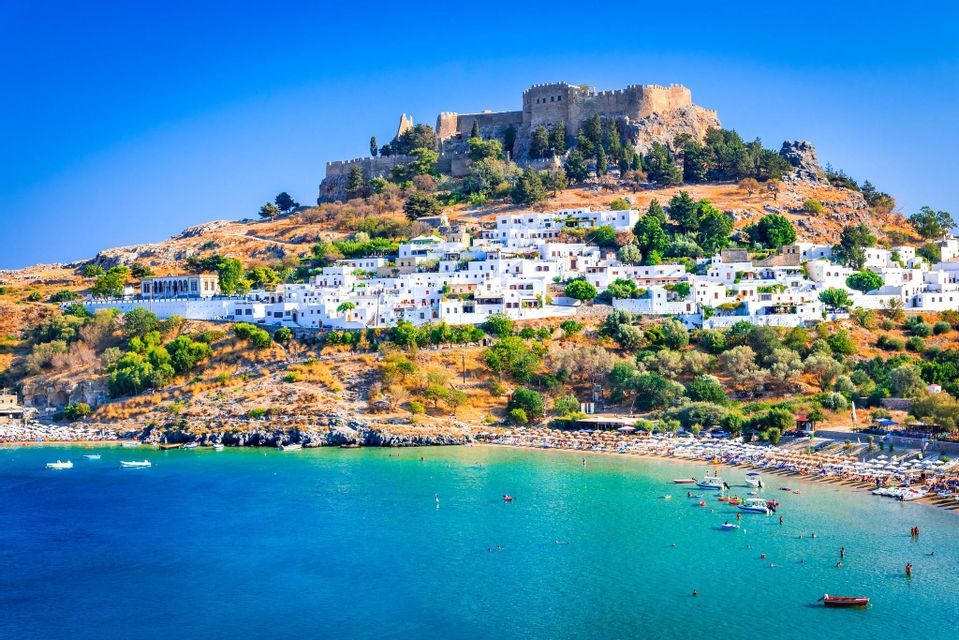 Un'antica acropoli sorge sopra un villaggio di case bianche, affacciato su una baia turchese con una spiaggia affollata in una giornata di sole.