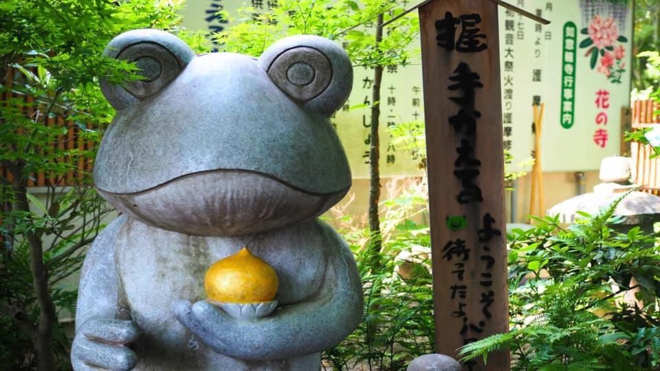 Una statua in pietra di una rana che tiene un piccolo oggetto dorato in un giardino giapponese, circondata da una ricca vegetazione verde.