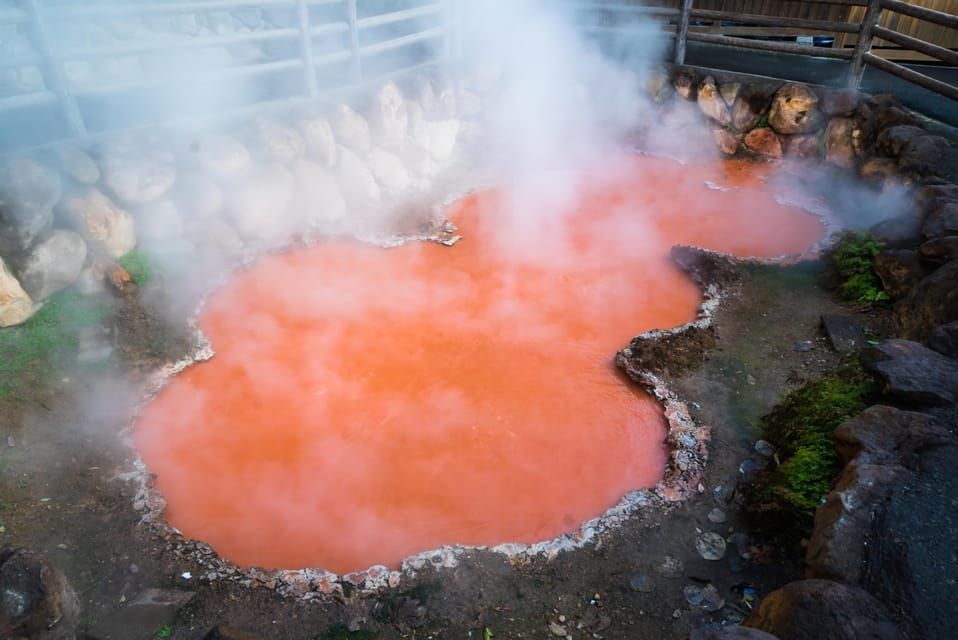 Una pozza d'acqua fumante, rosso-arancione, è circondata da rocce scure e una bassa recinzione di legno.
