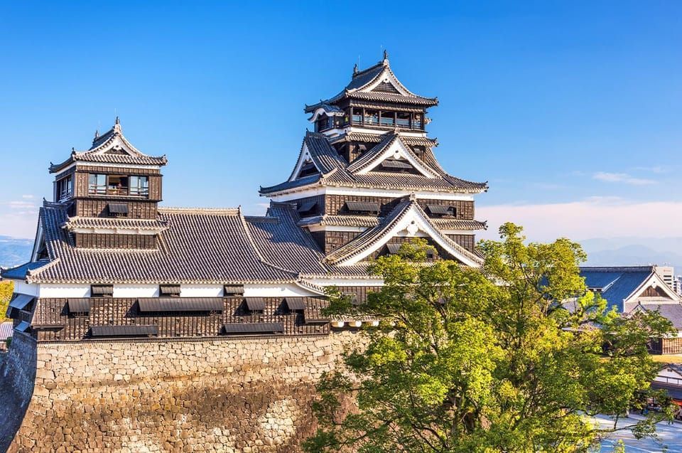 Un castello giapponese a più livelli con legno scuro e tetti di tegole si erge su un'alta cinta muraria di pietra sotto un cielo azzurro e limpido.