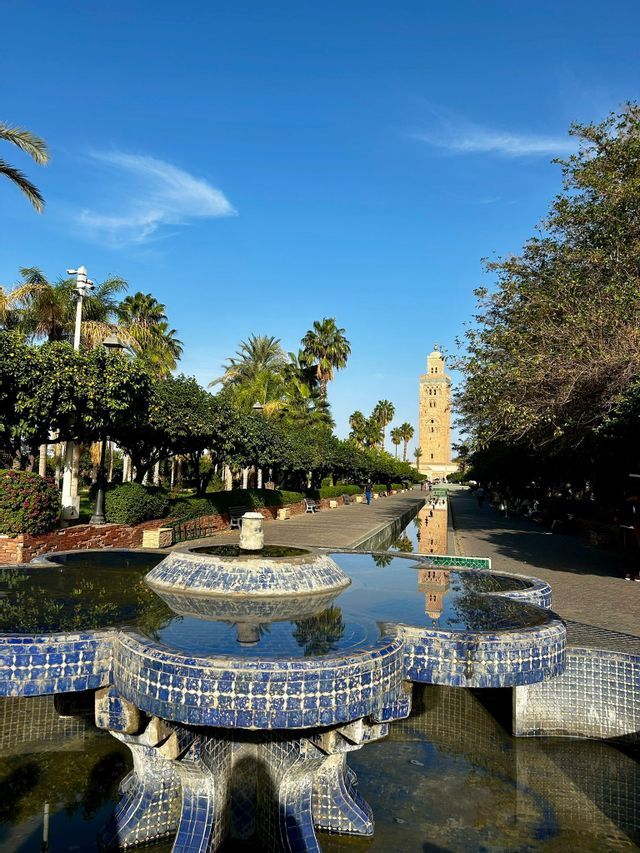 Una fuente de azulejos azules en un parque soleado con su agua reflejando una torre alta y palmeras contra un cielo azul claro.