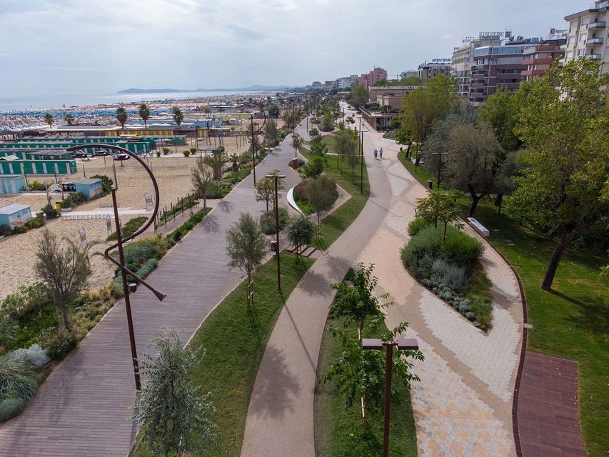 Veduta aerea di un lungomare paesaggistico con vialetti sinuosi, adiacente a una spiaggia sabbiosa e una fila di edifici.