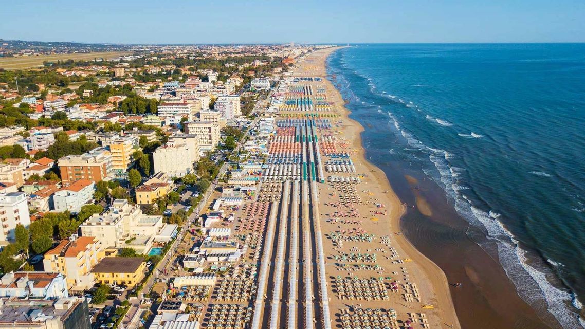 Una vista aerea di una lunga costa sabbiosa con file di ombrelloni colorati accanto a una città costiera e l'oceano.