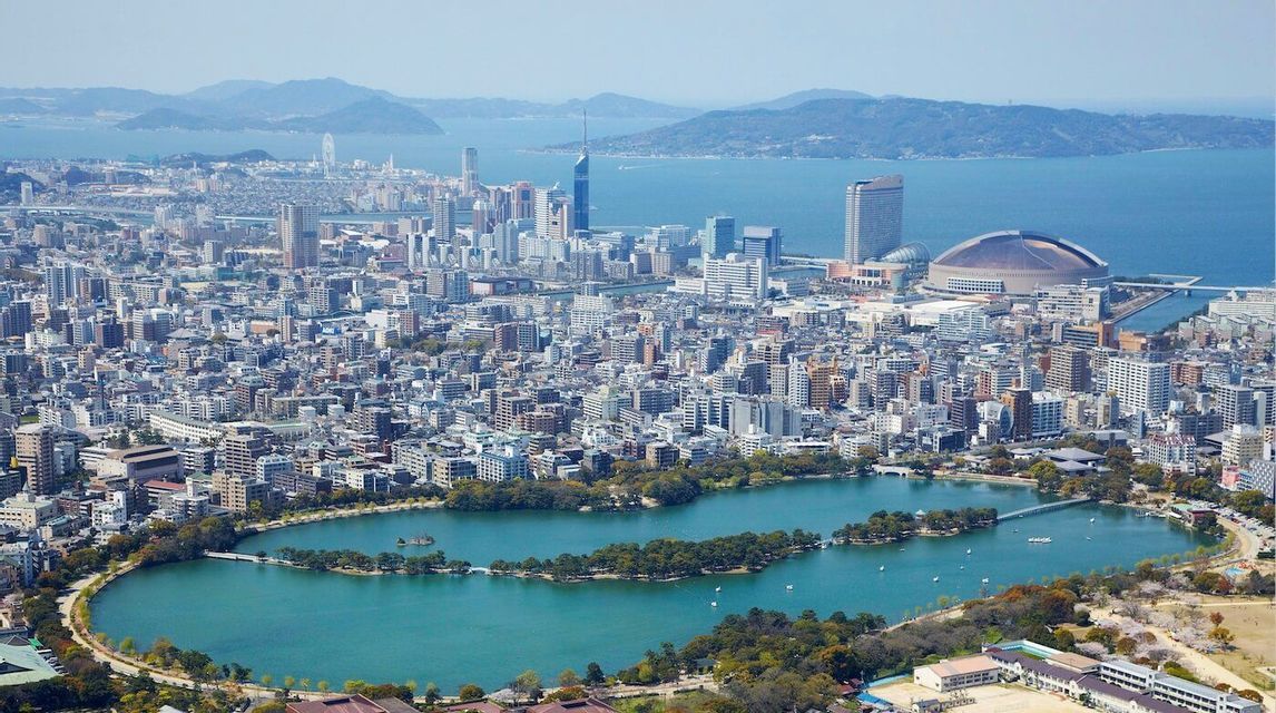 Una vista aerea di una vasta città costiera con un grande lago serpeggiante in un parco, moderni grattacieli e uno stadio a cupola sul mare.