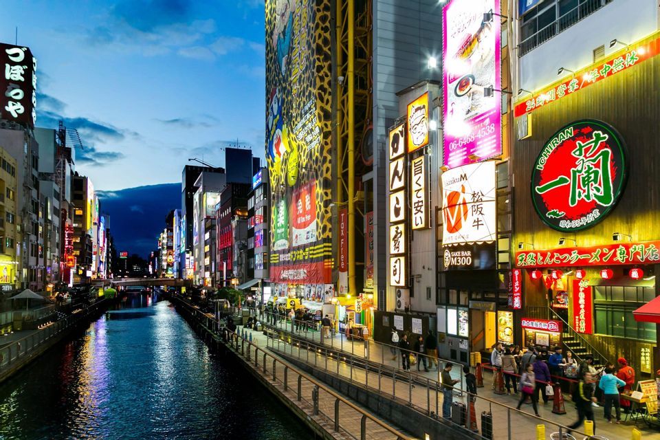 Un canale urbano al tramonto, con persone che passeggiano su un lungofiume fiancheggiato da edifici illuminati da insegne al neon.