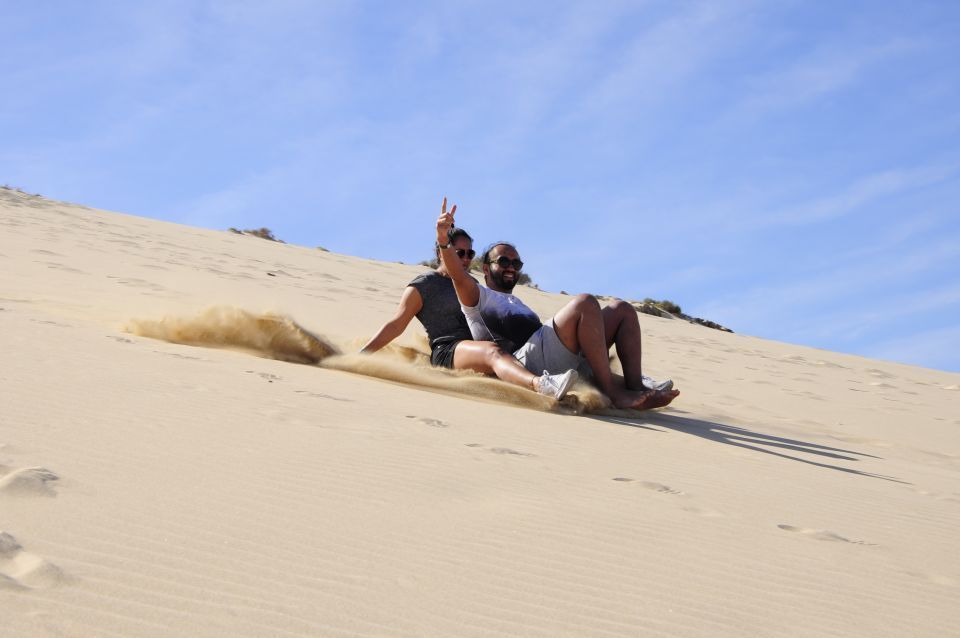 Un hombre y una mujer en un viaje en grupo de WeRoad se deslizan por una gran duna de arena, levantando arena bajo un cielo soleado.