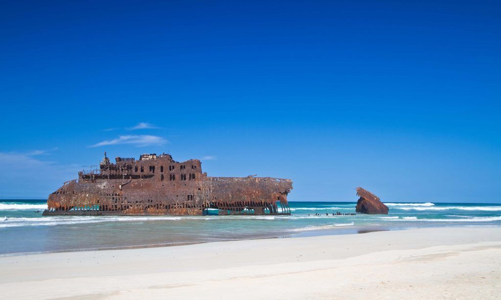 Un gran naufragio oxidado descansa varado en una playa de arena blanca con agua turquesa bajo un cielo azul claro.