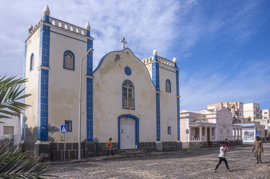 La fachada de una iglesia blanca con ribetes azules en una calle adoquinada, con gente pasando bajo un cielo azul nublado.