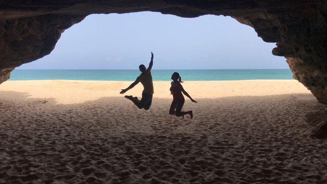 Siluetas de dos personas de un viaje en grupo de WeRoad saltando en una playa, enmarcadas por la entrada de una cueva.