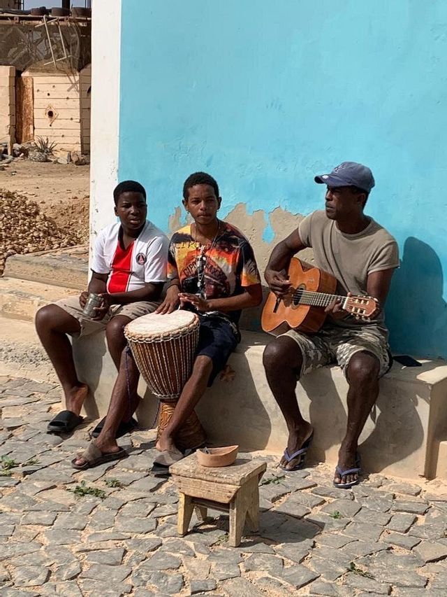 Tres hombres sentados en un saliente de piedra tocando un djembe y una guitarra acústica en una calle empedrada.