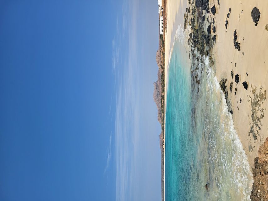 Una playa de arena con rocas volcánicas negras se encuentra con el océano turquesa claro, con colinas distantes bajo un vasto cielo azul.