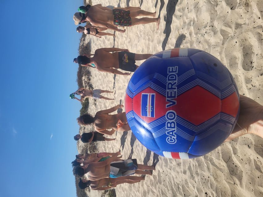 Una mano sostiene un balón de fútbol de Cabo Verde en una playa, con un viaje en grupo de WeRoad de fondo.