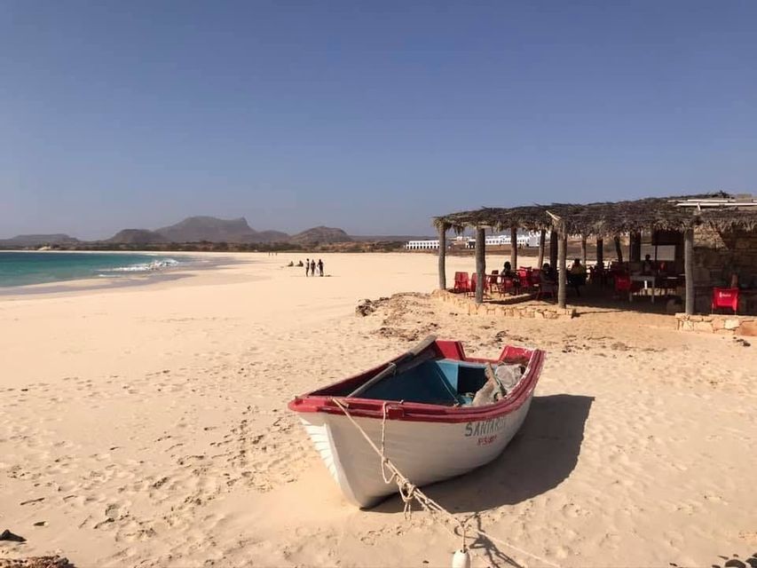 Un barco blanco y rojo descansa sobre una vasta playa de arena junto a un chiringuito con techo de paja, con montañas al fondo.