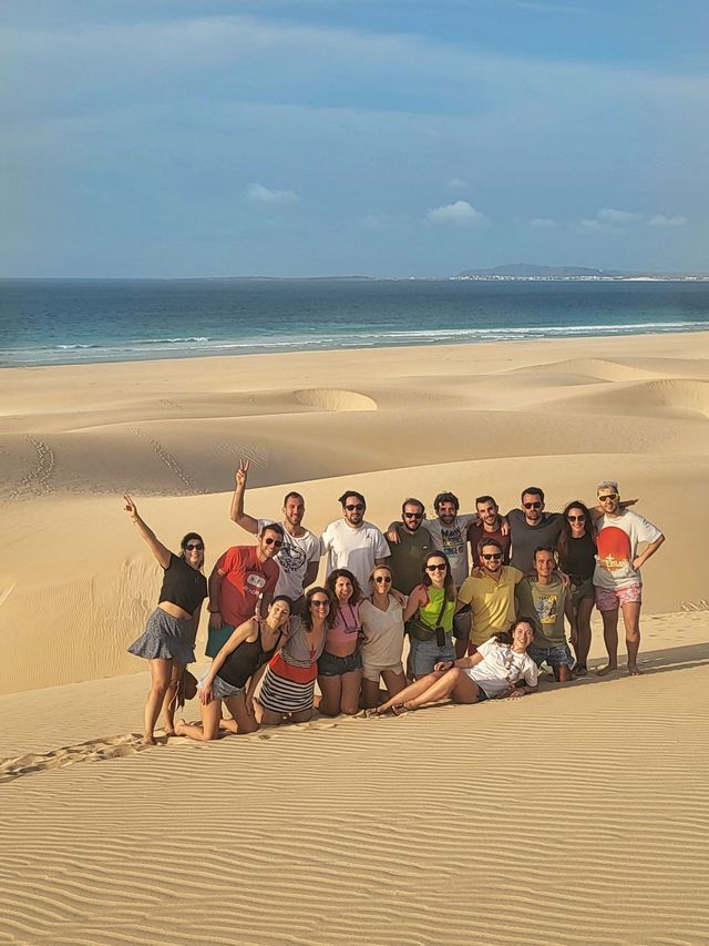 Un viaje en grupo de WeRoad posando para una foto en grandes dunas de arena con el océano y una costa distante al fondo.