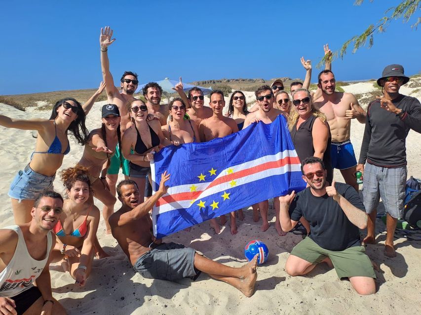 Un grupo de WeRoad posa en una playa de arena bajo un cielo azul claro, sosteniendo la bandera nacional de Cabo Verde.