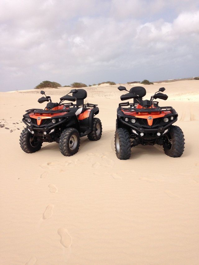 Dos quads naranjas y negros estacionados en vastas dunas de arena con vegetación escasa bajo un cielo nublado.