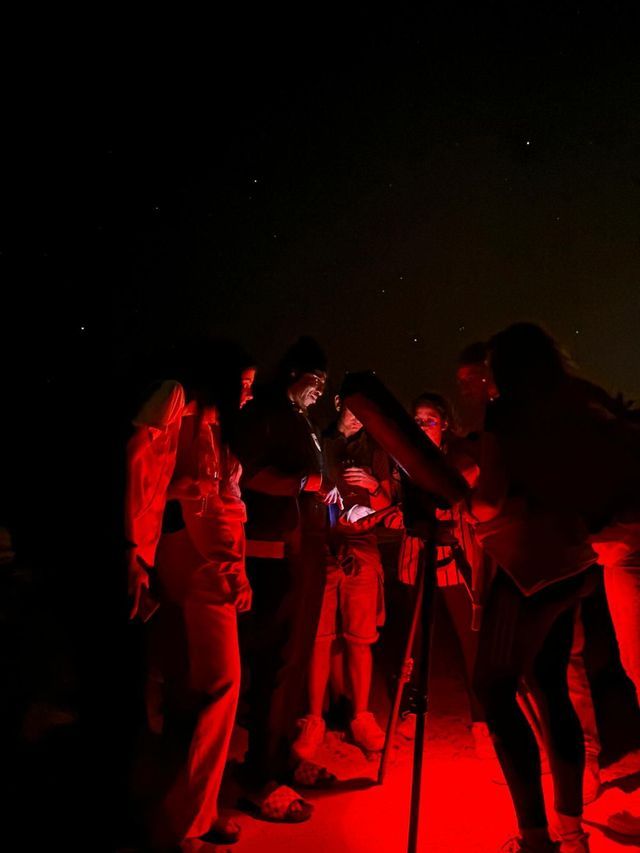 Un viaje en grupo de WeRoad se reúne alrededor de un telescopio para observar las estrellas por la noche, iluminado por una luz roja.