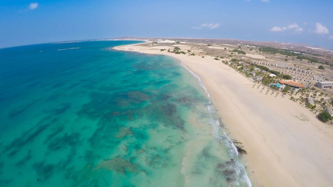 Vista aérea de una amplia playa de arena bordeando un océano turquesa con arrecifes submarinos visibles bajo un cielo azul.