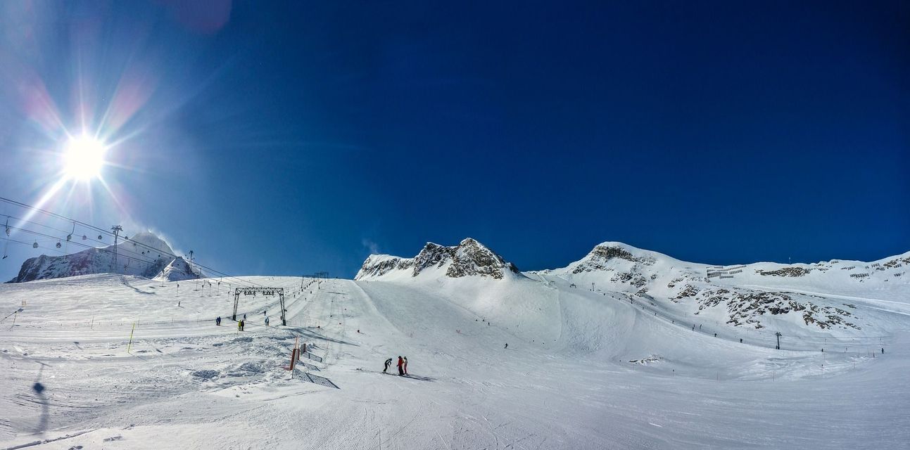 Un ampio pendio montano innevato con persone che sciano sotto un sole splendente e un cielo azzurro chiaro.