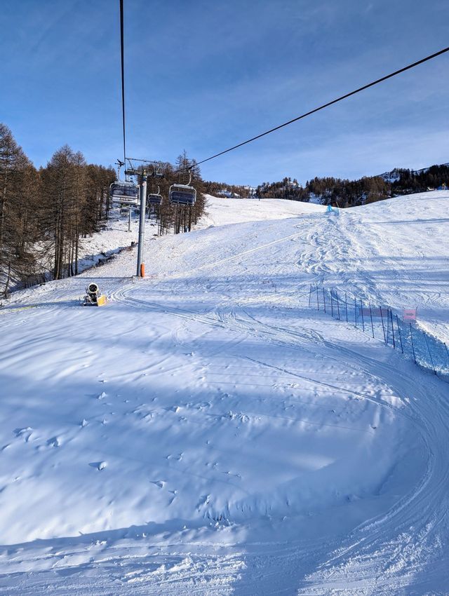 Una vista da una seggiovia che domina un'ampia pista da sci innevata in una giornata di sole con un cielo azzurro limpido.