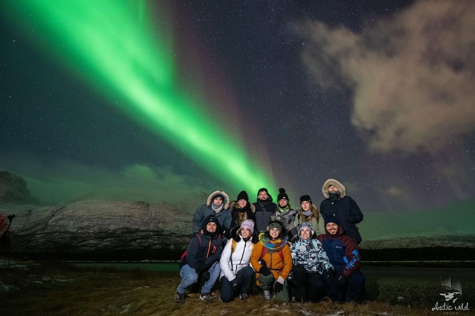 Un viaje en grupo de WeRoad con ropa de invierno posando para una foto bajo una aurora boreal verde vibrante y un cielo nocturno estrellado.