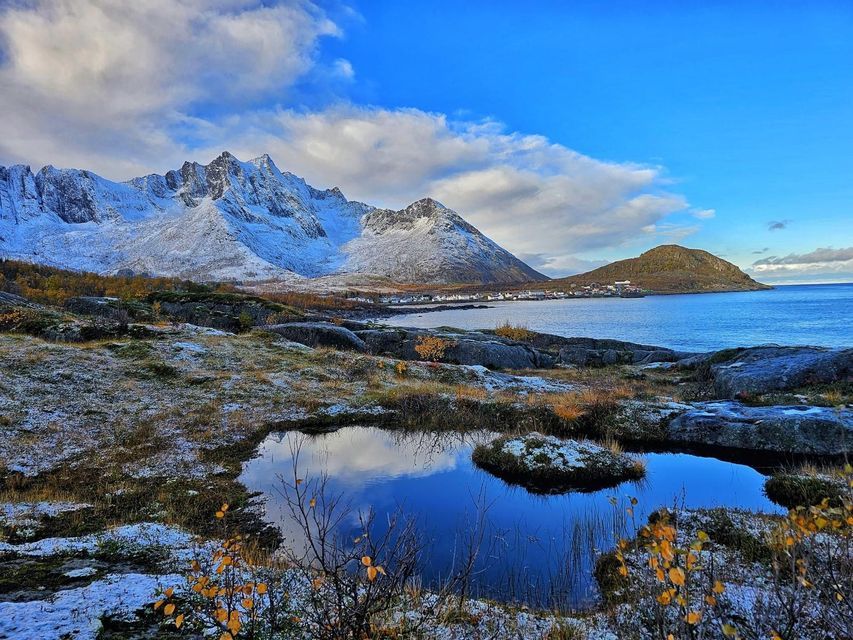 Un paisaje nevado con un estanque que refleja el cielo azul, con vistas a un pueblo costero enclavado junto a montañas cubiertas de nieve.