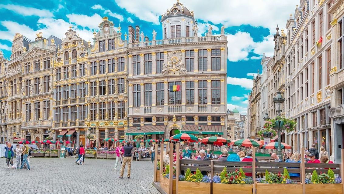 Una amplia plaza adoquinada llena de gente en cafés al aire libre, rodeada de edificios ornamentados con detalles dorados bajo un cielo azul.