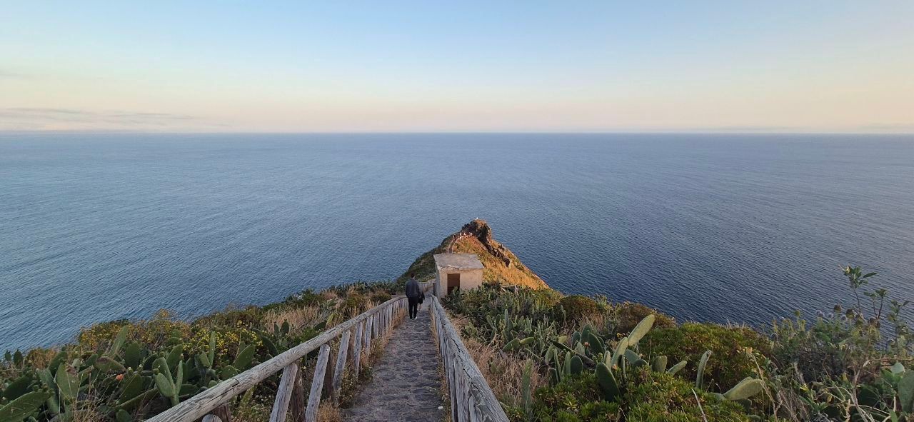 Una persona cammina lungo un sentiero in pietra con un parapetto in legno su un promontorio costiero che si affaccia su un vasto mare al tramonto.