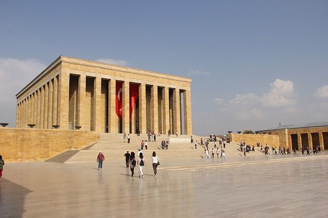 Turisti attraversano una grande piazza e salgono i gradini di un imponente edificio in pietra con colonne e una bandiera turca.