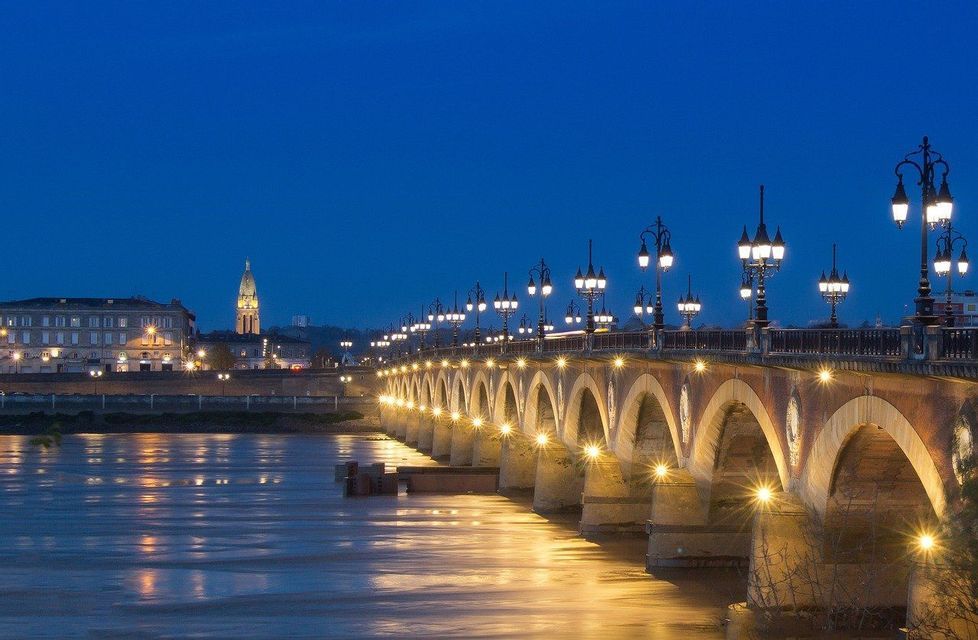 Un puente de piedra iluminado con muchos arcos y farolas cruza un amplio río al atardecer bajo un cielo azul profundo.