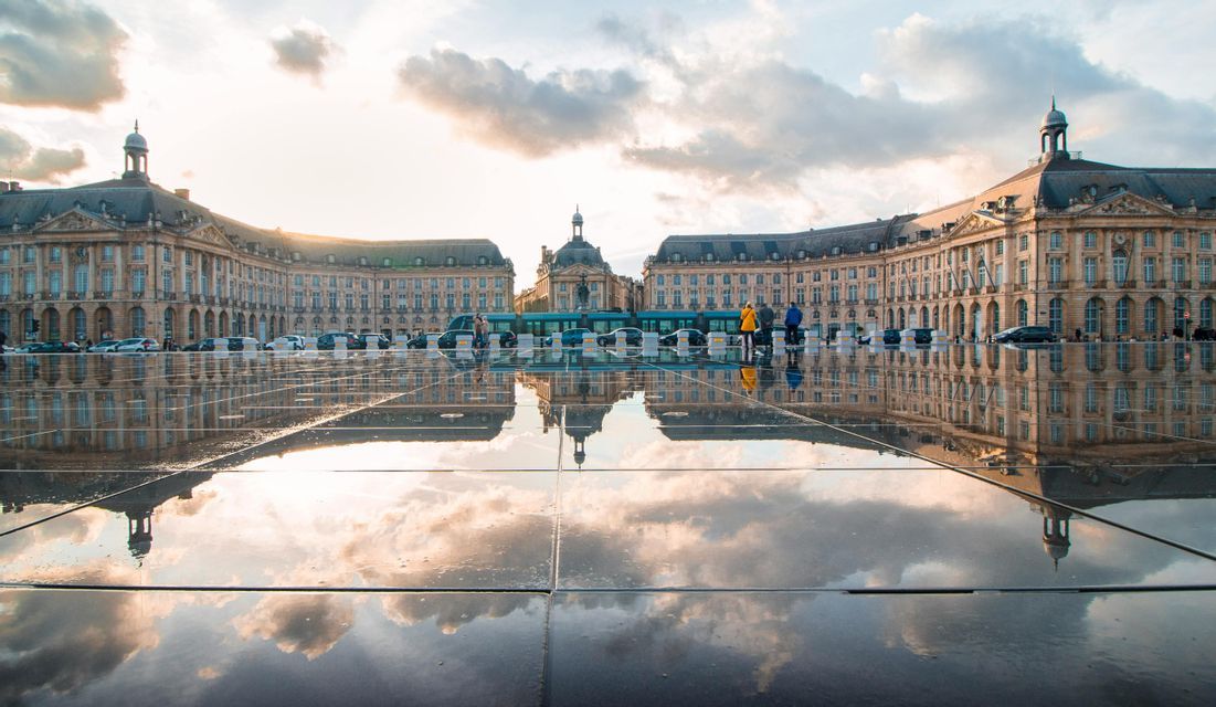 Un gran complejo de edificios clásicos y un cielo nublado se reflejan perfectamente en una plaza mojada al atardecer.