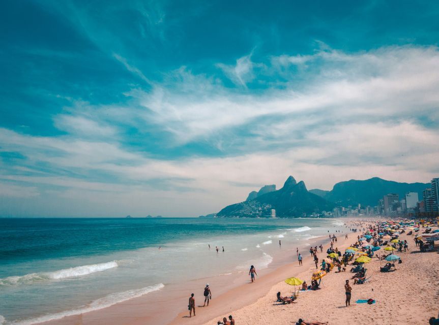 Une plage de sable animée, avec des baigneurs et des personnes prenant le soleil, une ville et des montagnes en toile de fond sous un ciel bleu éclatant.