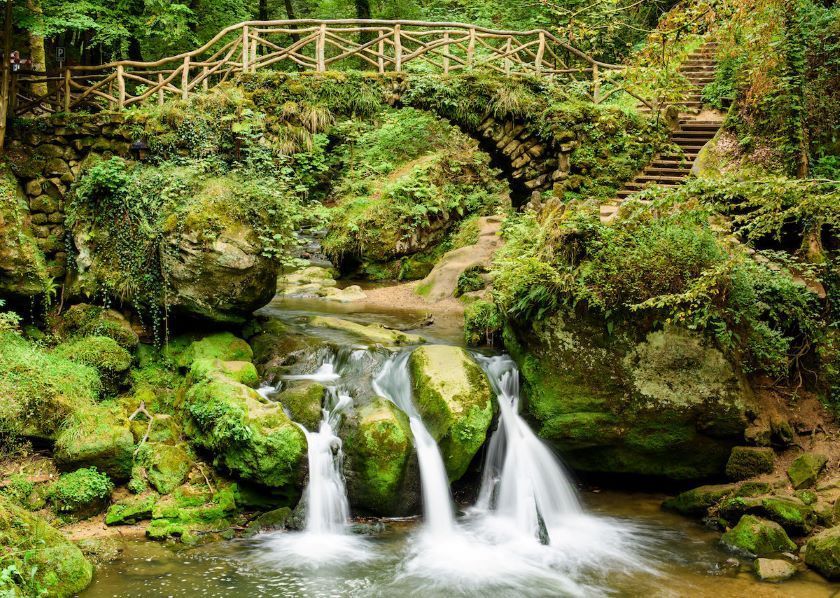 Un ponte di pietra muschioso con una ringhiera di legno si inarca su un piccolo ruscello con cascate in una foresta lussureggiante e verde.
