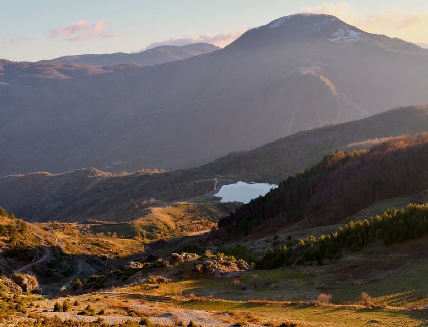 Un piccolo lago è immerso in una vasta valle di montagna con dolci colline, illuminato dalla calda luce del tramonto.